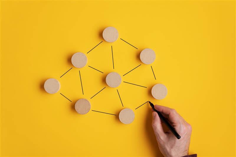 A hand draws connections between wooden dots on a yellow background using a black marker.