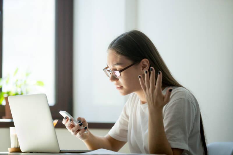 A young woman sitting at a desk looks annoyed at her phone, with one hand thrown up in frustration