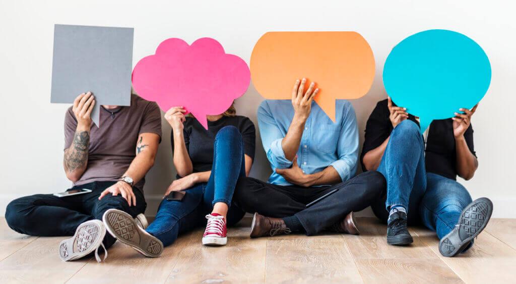 Four young-looking people sit closely and casually on the floor, holding paper cutouts shaped like speech bubbles and thought bubbles in front of their faces.