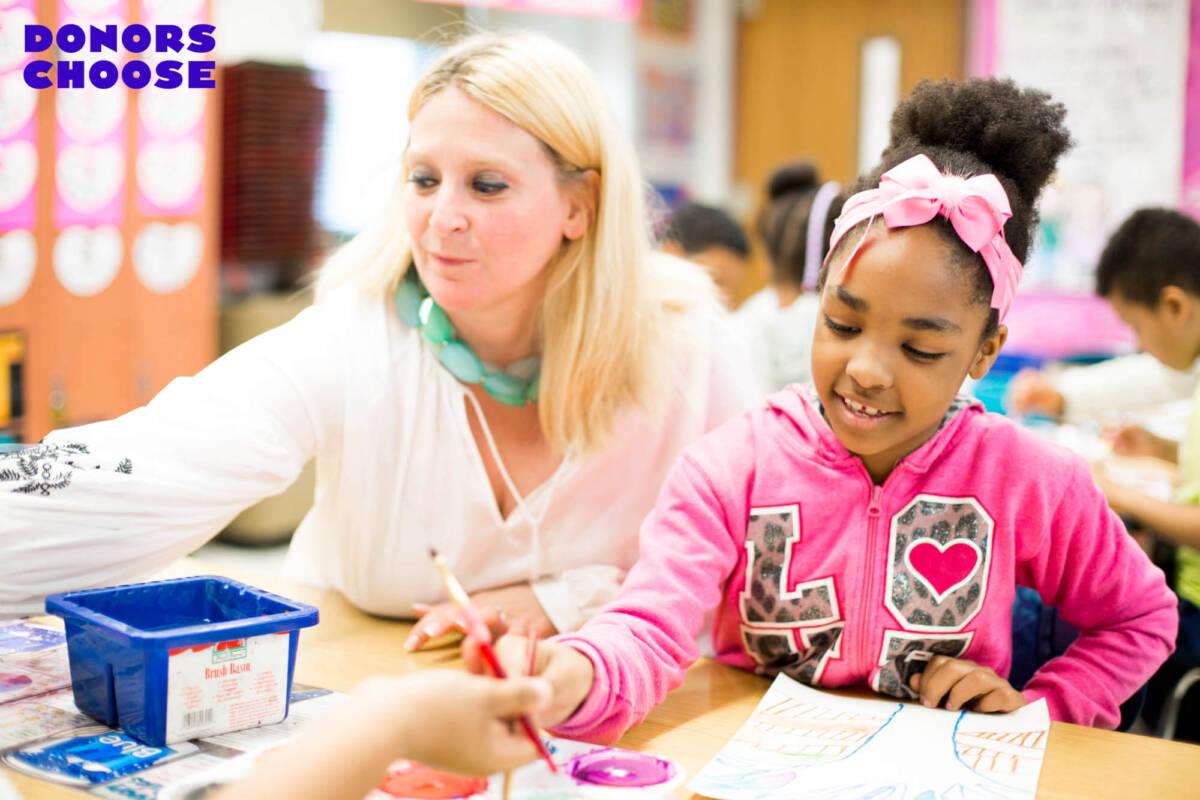 Blonde-haired teacher works with young students, blue DonorsChoose logo superimposed overhead