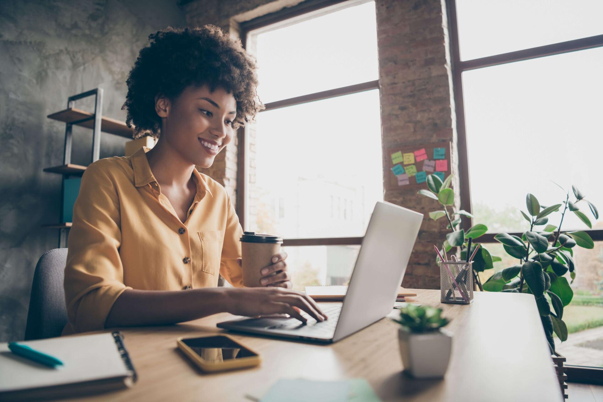 A smiling young woman looks at her computer with one hand on the keyboard and the other holding a paper cup