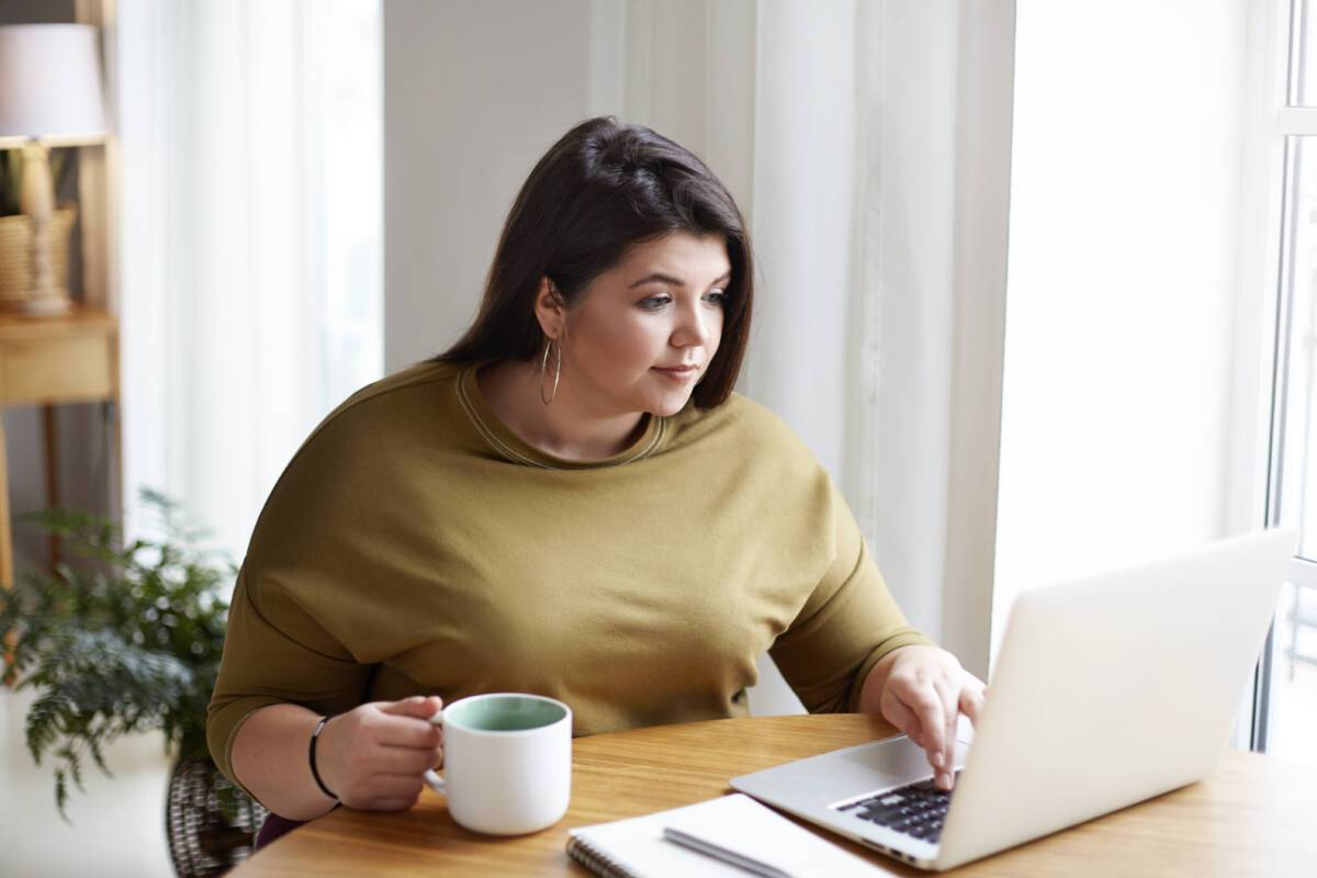 Woman in yellow sweater working in front of open laptop, sitting in home office interior, drinking coffee