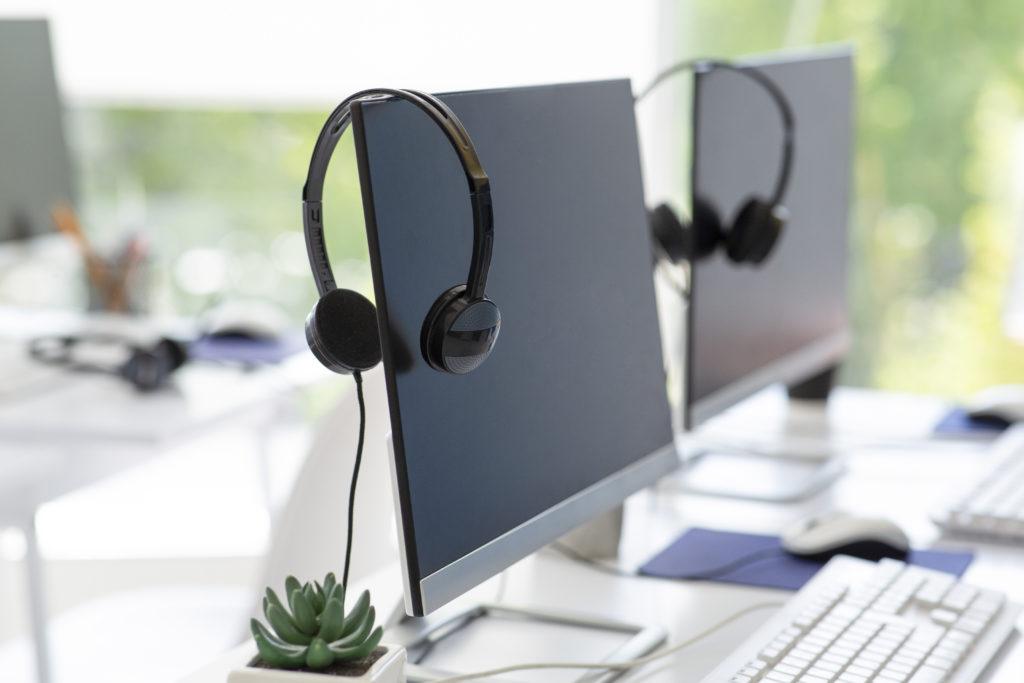 Headsets hang on two powered-off desktop monitors in an occupied office space illuminated by bright, natural light