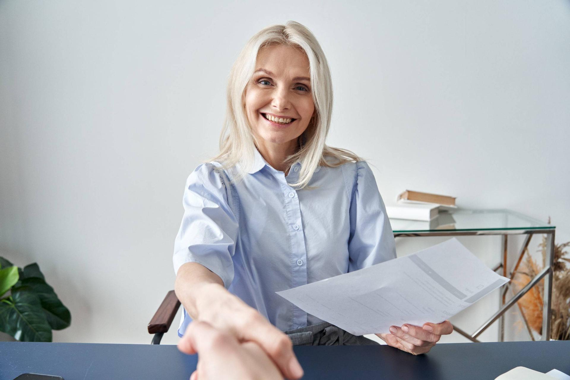 Person with white hair smiles and shakes hand of another person on opposite side of the camera