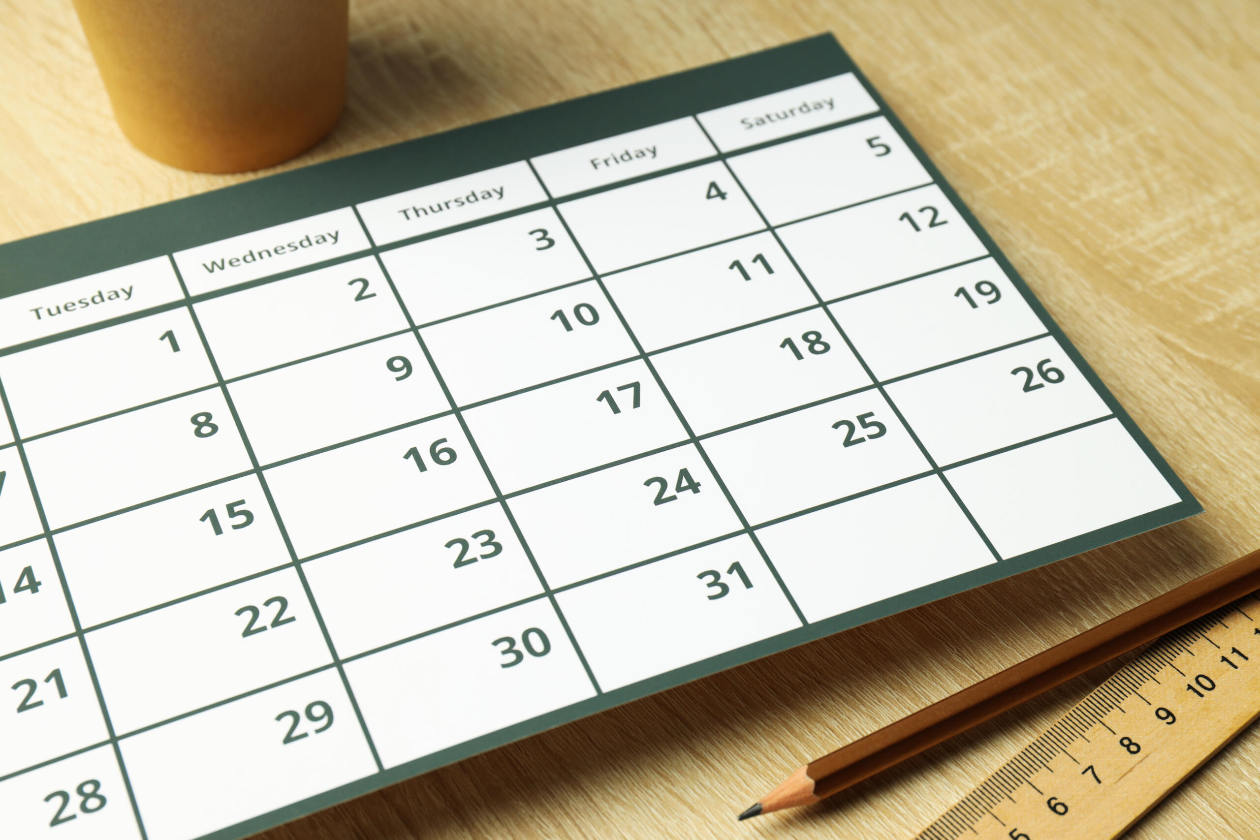 Effortless scheduling for your business: close-up of calendar on wooden surface with pencil, ruler, and coffee cup partially in frame