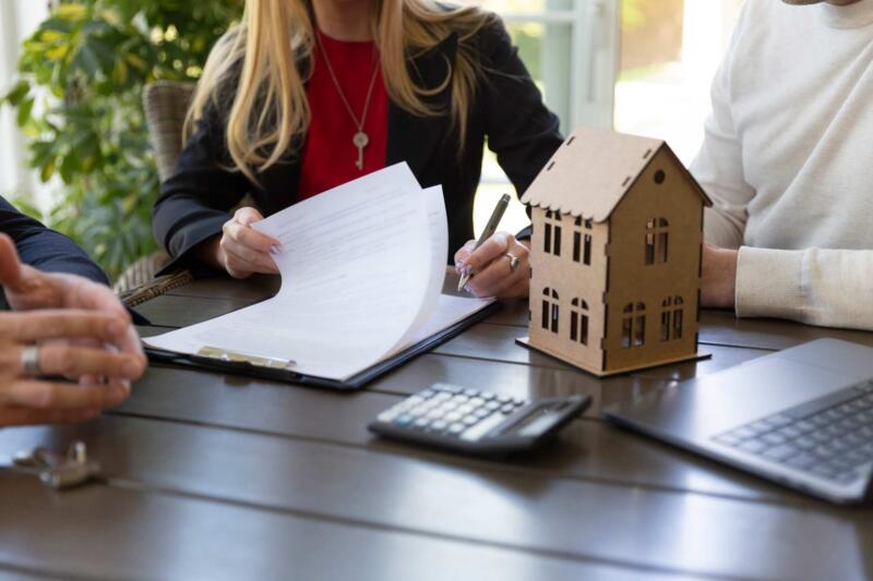 A collection of people filling out paperwork while sitting around a table. A small wooden house is on the table next to a calculator and a laptop.