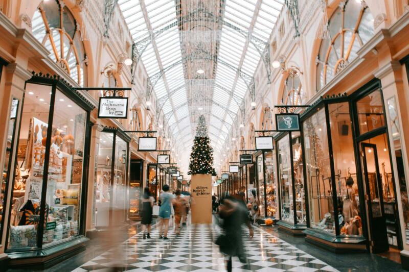 A photo of a busy shopping mall during the holiday season. The mall has shoppers bustling back and forth and in the center of the mall there is a Christmas tree. This image was taken by Heidi Fin