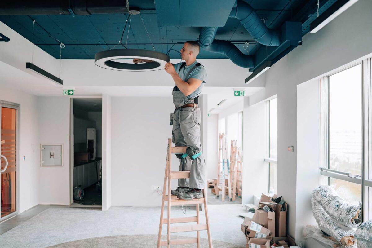 A shot of an electrician on a ladder installing an overhead circular light fixture.