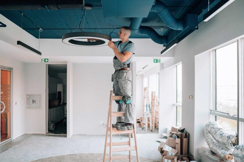 A shot of an electrician on a ladder installing an overhead circular light fixture.