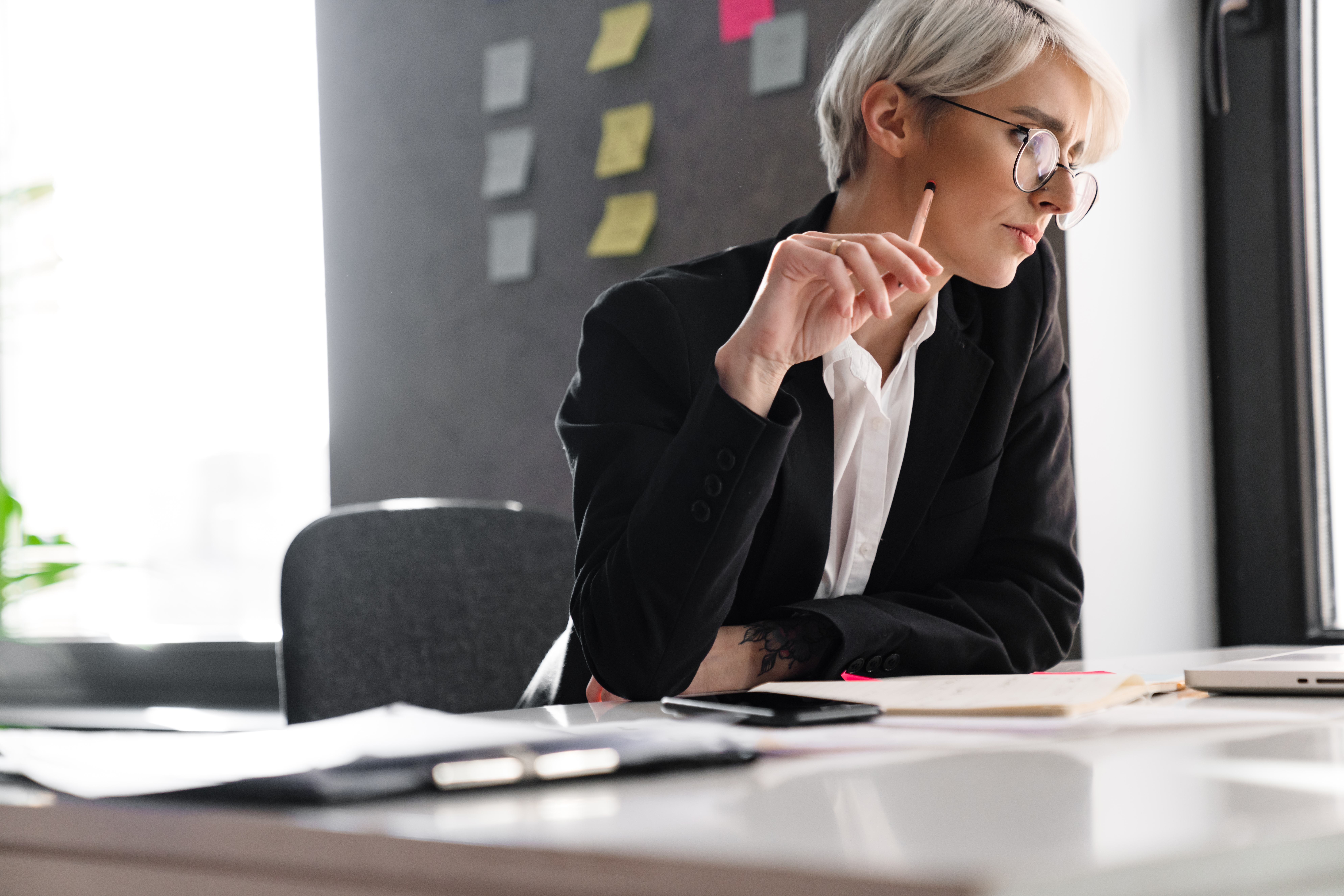 Is AI working for or against your firm: A professional with short white hair sits at a desk in a well-lit room with a gray wall covered in sticky notes, staring intently at something offscreen.