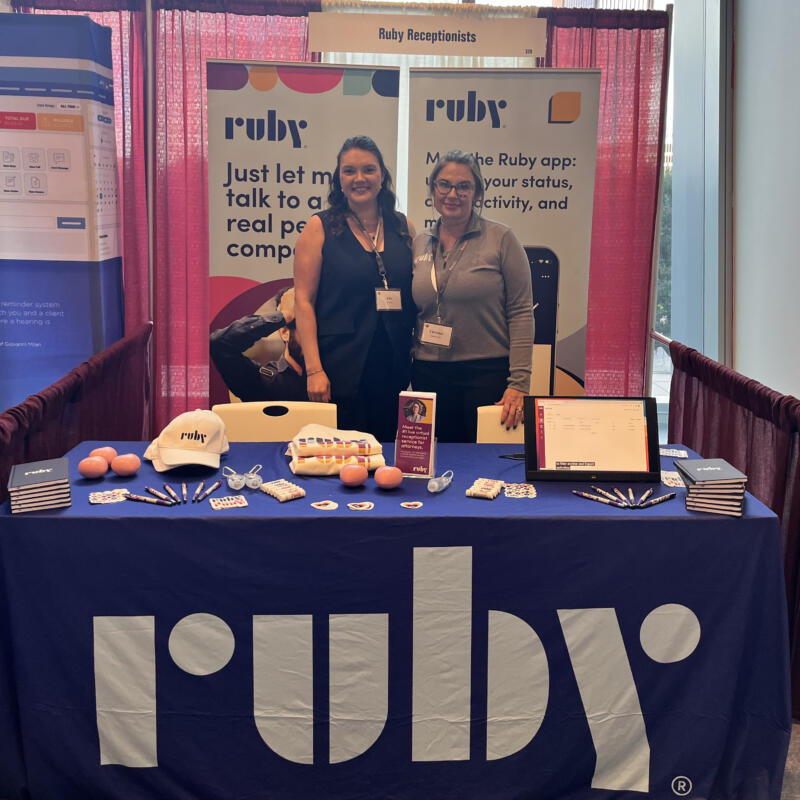 Ellie Miller and Christina Urban from Ruby stand behind a colorful Ruby table at the State Bar of Texas Annual Meeting in Houston. The table is topped with branded notebooks, hats, pens, stress balls, and other Ruby swag, along with a tablet showing the Ruby dashboard. Behind Ellie and Tina are two standee signs reading "Just let me talk to a real person company." and "Meet the Ruby app: update your status, check activity, and more."
