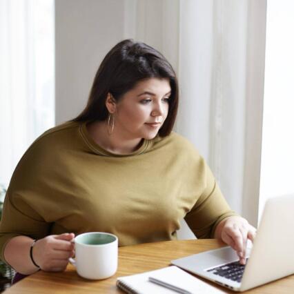 Woman in yellow sweater working in front of open laptop, sitting in home office interior, drinking coffee