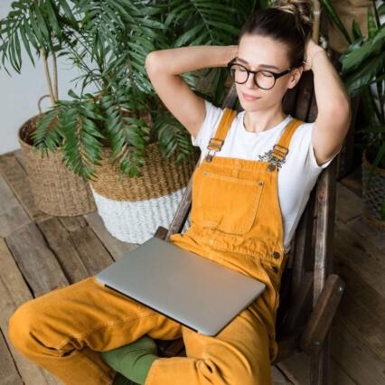 A woman in yellow overalls sits on a hardwood floor among houseplants with a closed laptop, her hands behind her head
